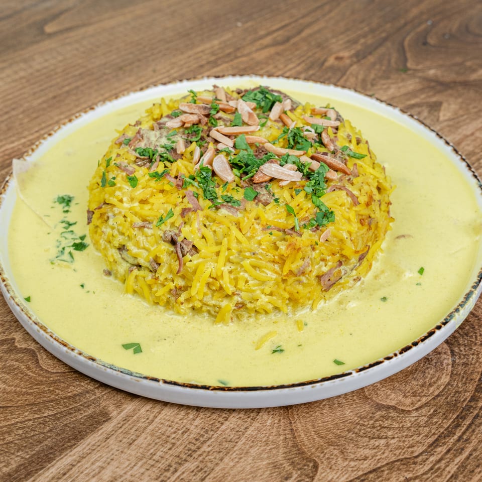 Yellow rice dish topped with sliced almonds, chopped herbs, and meat, surrounded by creamy sauce on a white plate.