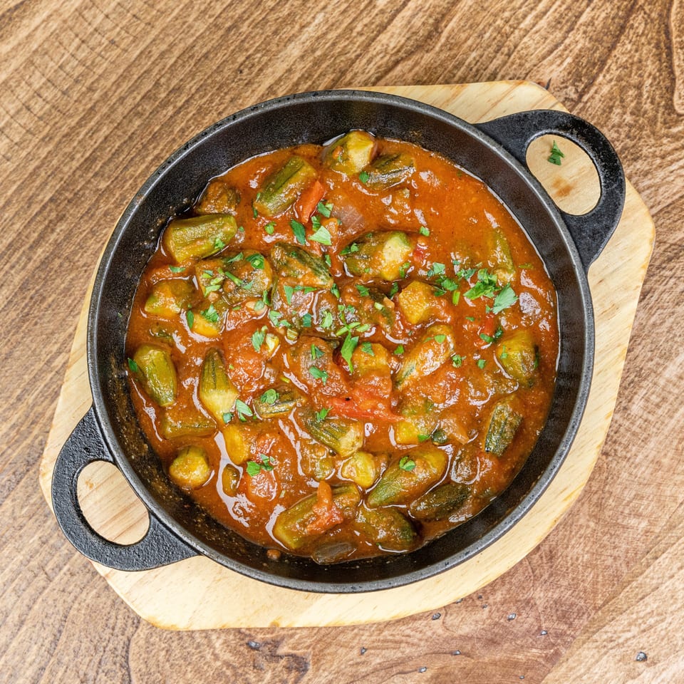 Cast-iron skillet of okra and vegetables in rich tomato sauce, garnished with fresh herbs on a wooden serving board.