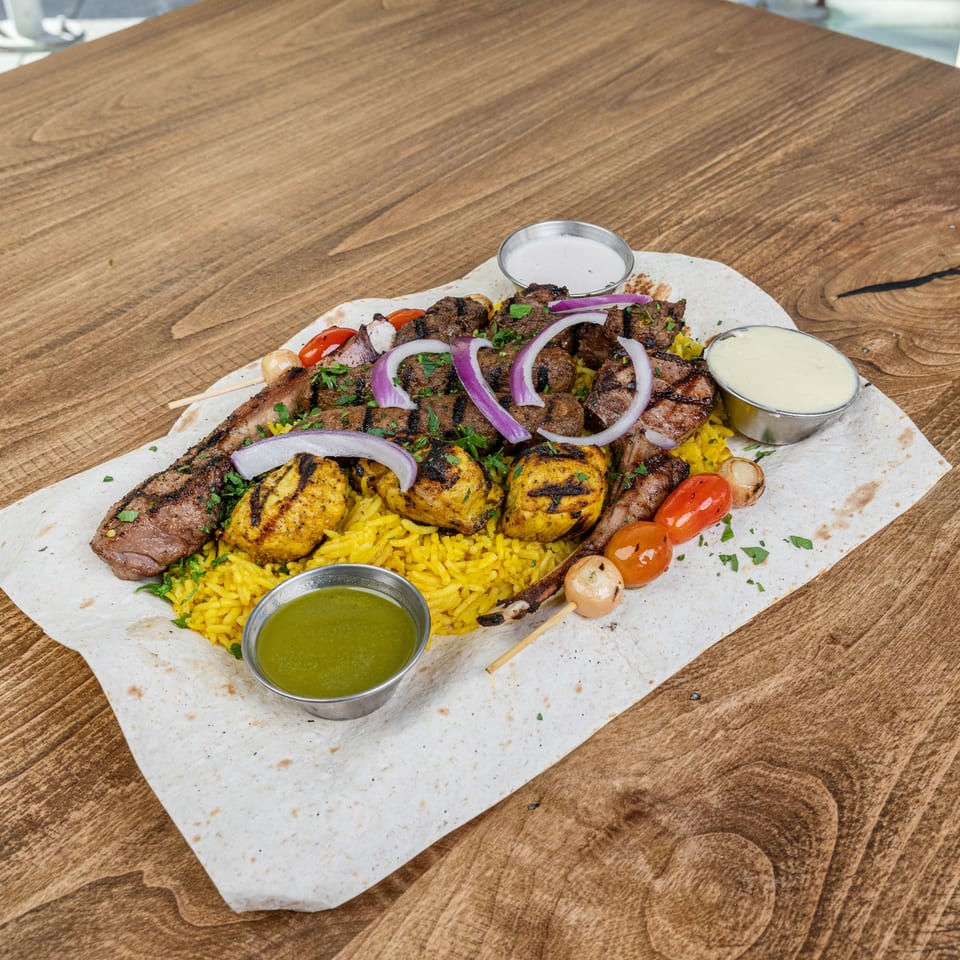 Assorted grilled meat and chicken skewers with yellow rice, vegetables, red onion, and dipping sauces served on flatbread.