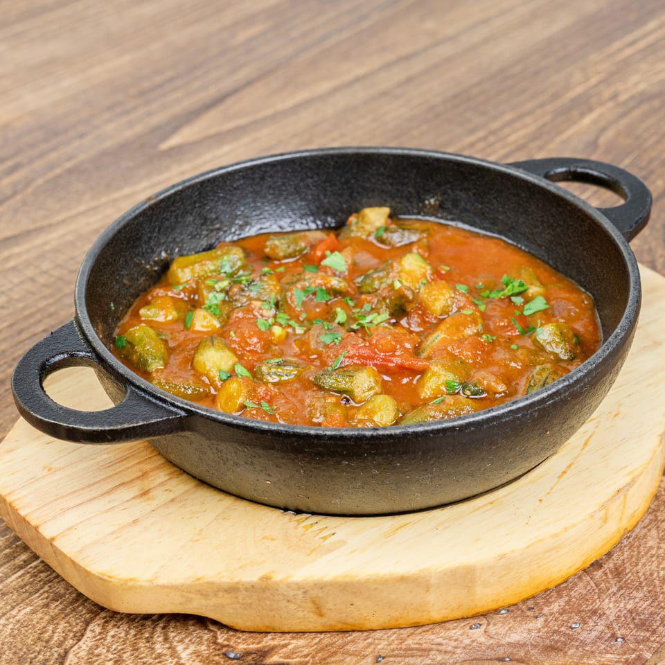 Cast-iron skillet filled with tomato-based vegetable stew, garnished with fresh herbs, on a wooden serving board.