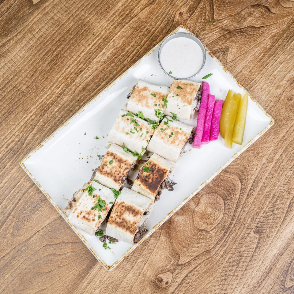 Grilled meat-filled flatbread pieces garnished with herbs, served with pickled vegetables and creamy dipping sauce on a white platter.