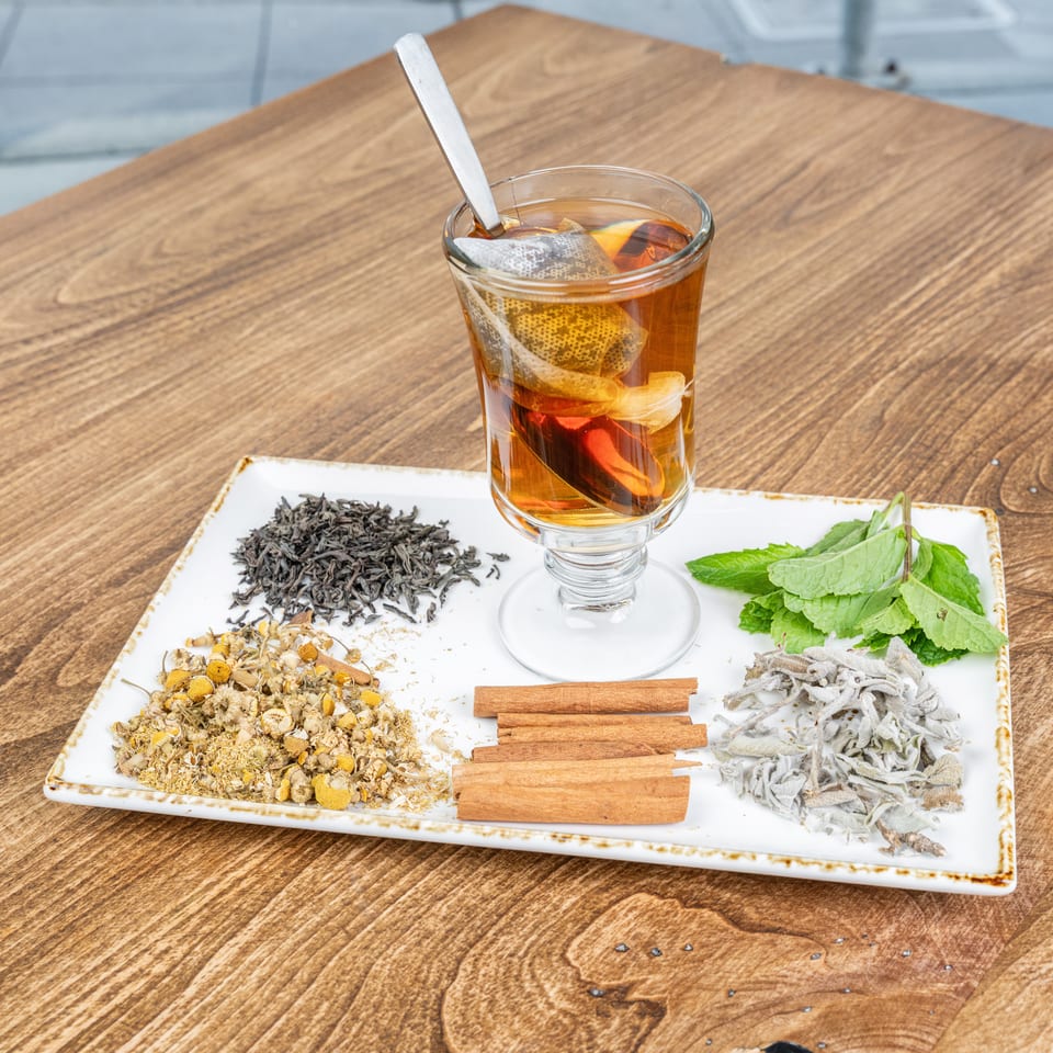 Glass of amber herbal tea with citrus slices and spoon, surrounded by loose herbs, mint, cinnamon, and sage on a tray.