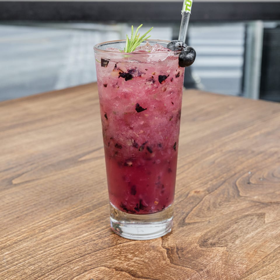 Pink berry cocktail with crushed ice, blueberries, rosemary garnish, and straw on a wooden restaurant table.
