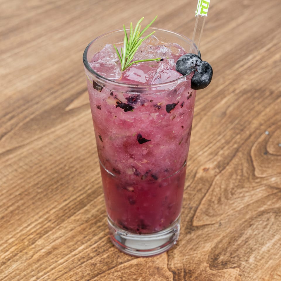 Pink berry cocktail with crushed ice, blueberries, rosemary garnish, and a straw on a wooden table.