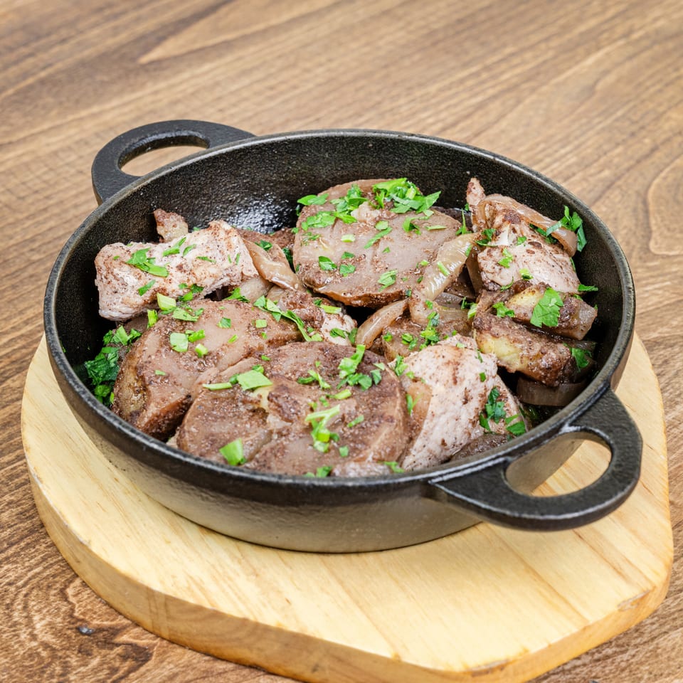 Skillet of seared meat pieces and onions garnished with fresh parsley on a wooden serving board.