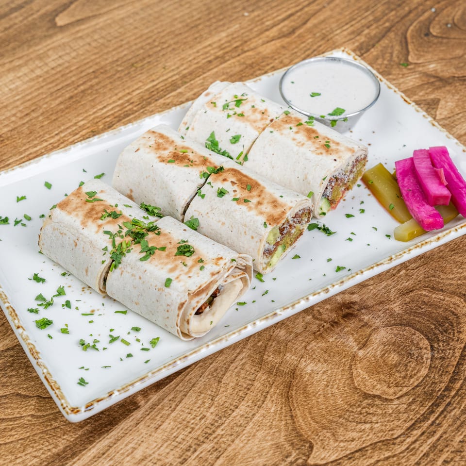 Grilled stuffed wraps garnished with parsley, served with creamy dipping sauce and pink and green pickles on a white platter.