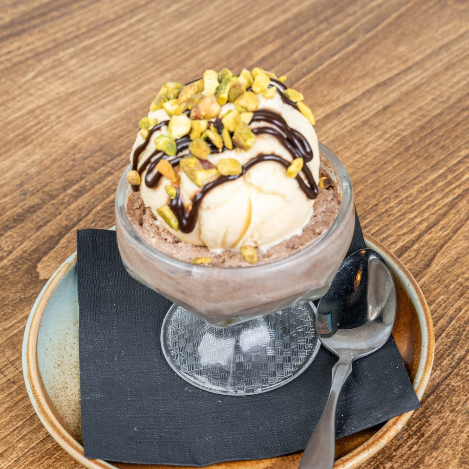 Chocolate dessert topped with vanilla ice cream, chocolate sauce, and pistachios, served in a glass dish with a spoon.