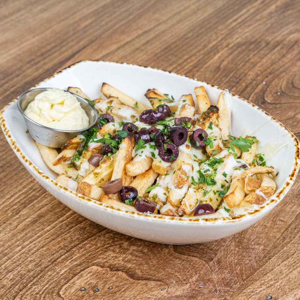 Loaded fries topped with melted cheese, grilled chicken, chopped herbs, and olives, served with creamy dipping sauce on a wooden table.